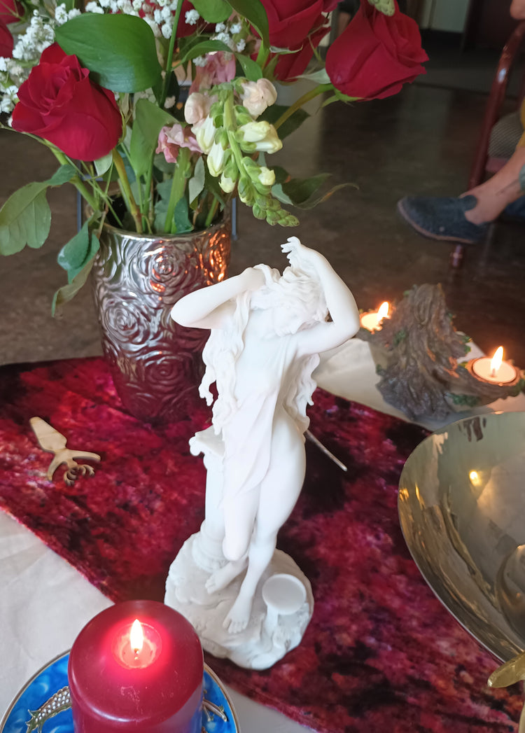 White statue of Aphrodite with a lit red candle and a vase of red roses in the background.