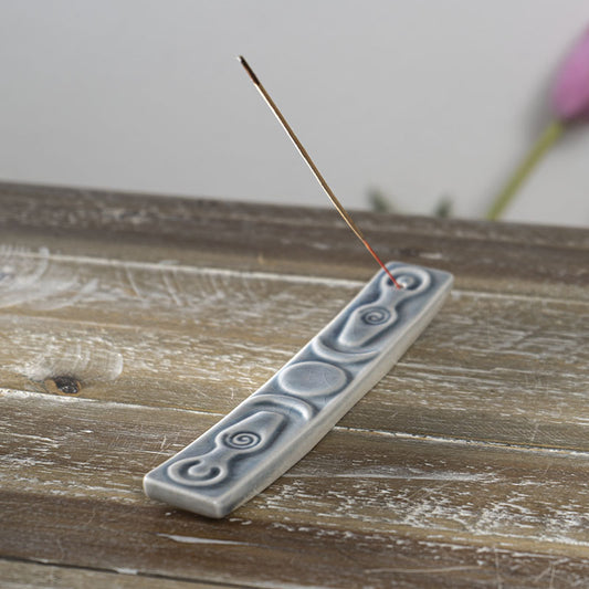 ceramic incense holder with images of a triple moon and two spiral goddesses. Shown with a stick of incense.