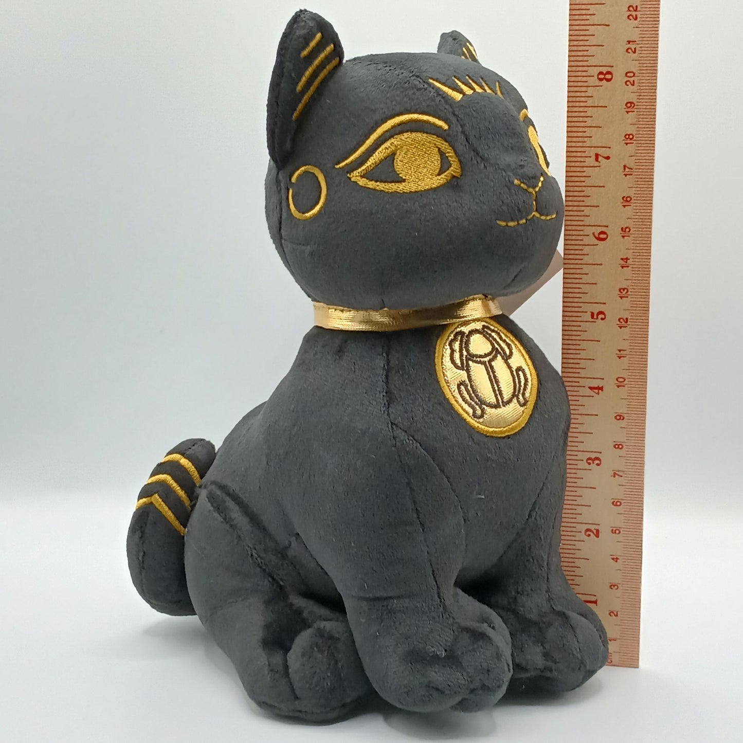Plush toy bastet cat with gold accents next to a ruler on a white background.  8+ inches tall
