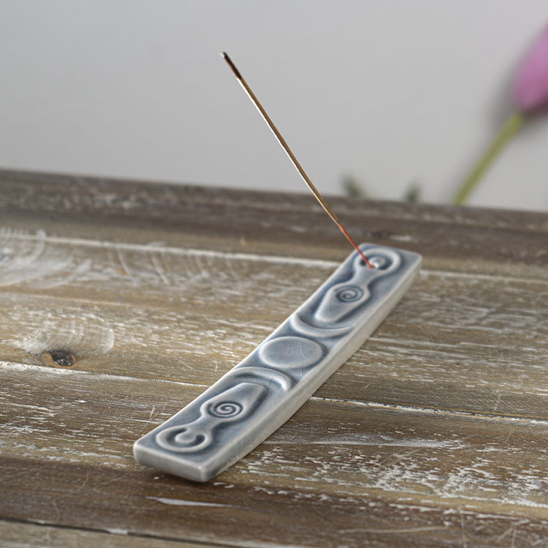 ceramic incense holder with images of a triple moon and two spiral goddesses. Shown with a stick of incense. 